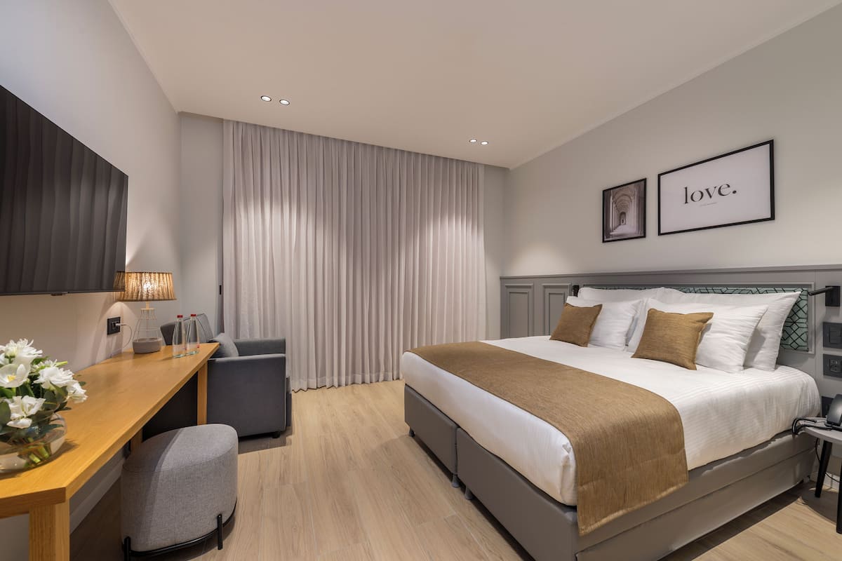 A modern hotel room with a large bed, a desk, and a chair. There are curtains, a lamp, and framed pictures on the wall.