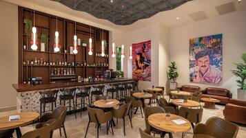 A modern bar area with a wooden counter, multiple seating options including round tables and chairs, and a ceiling with a geometric pattern.