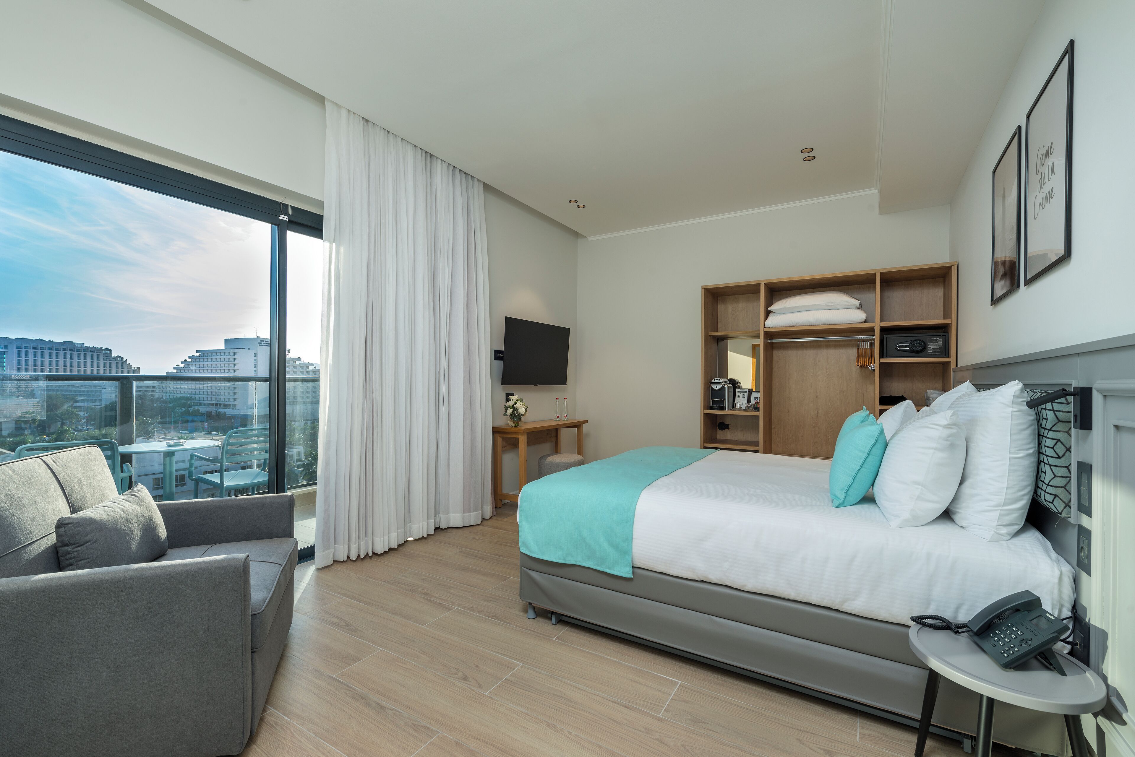Quarto de hotel moderno com uma cama grande, um sofá, uma escrivaninha e uma televisão. O quarto oferece vista para a cidade através de amplas janelas.