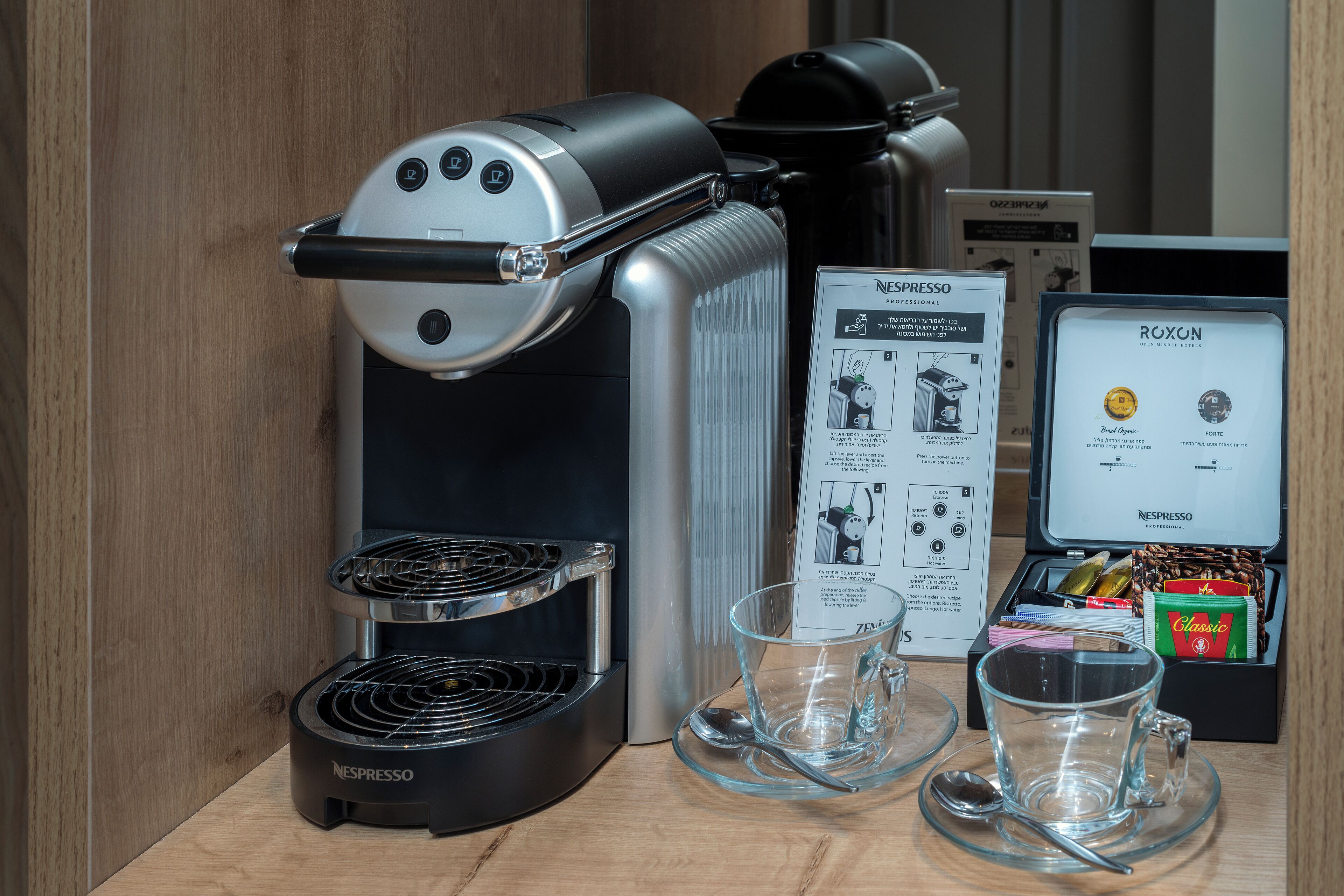 En Nespresso-kaffemaskin med en glaskanna och en kopp, en kaffeförpackning från Roxel och en sked på ett träbord.