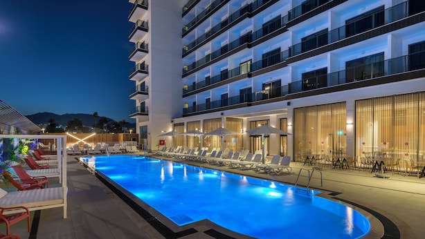 A swimming pool with lounge chairs and umbrellas, surrounded by a multi-story hotel building with balconies and large windows.