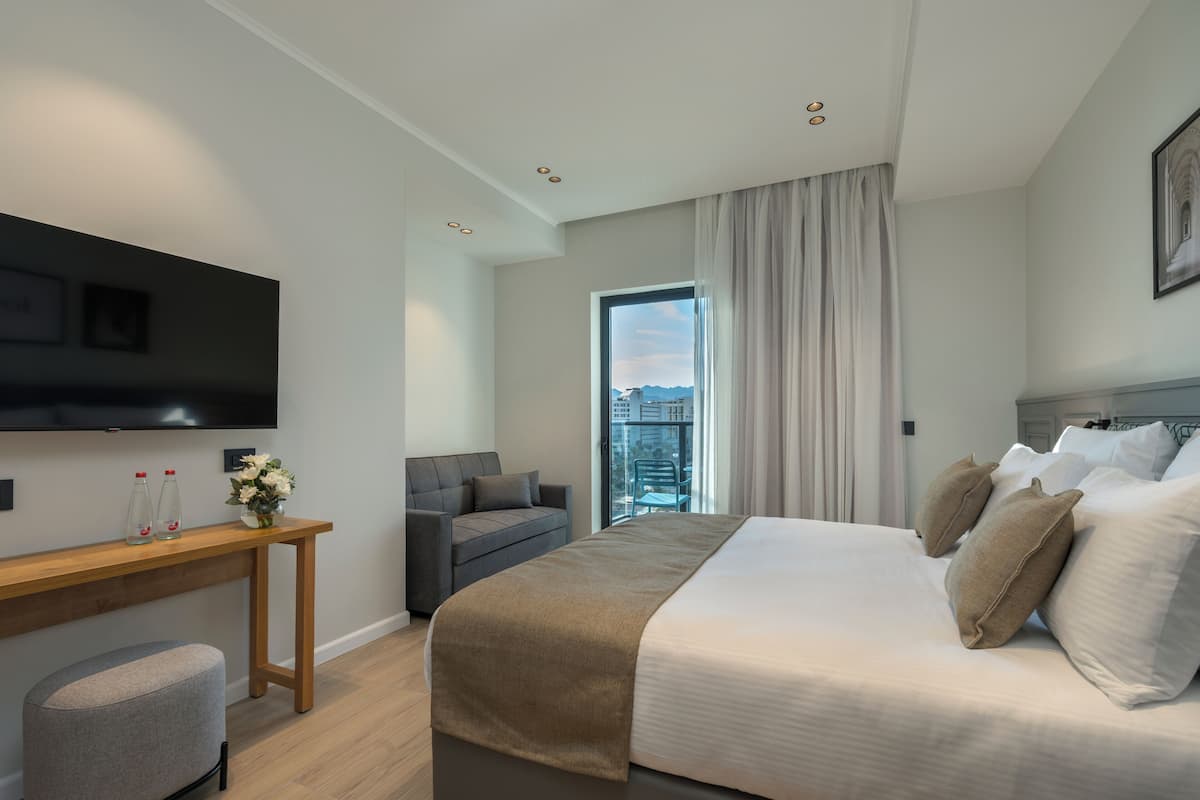 A modern hotel room with a large bed, a sofa, a TV mounted on the wall, and a window with a city view.