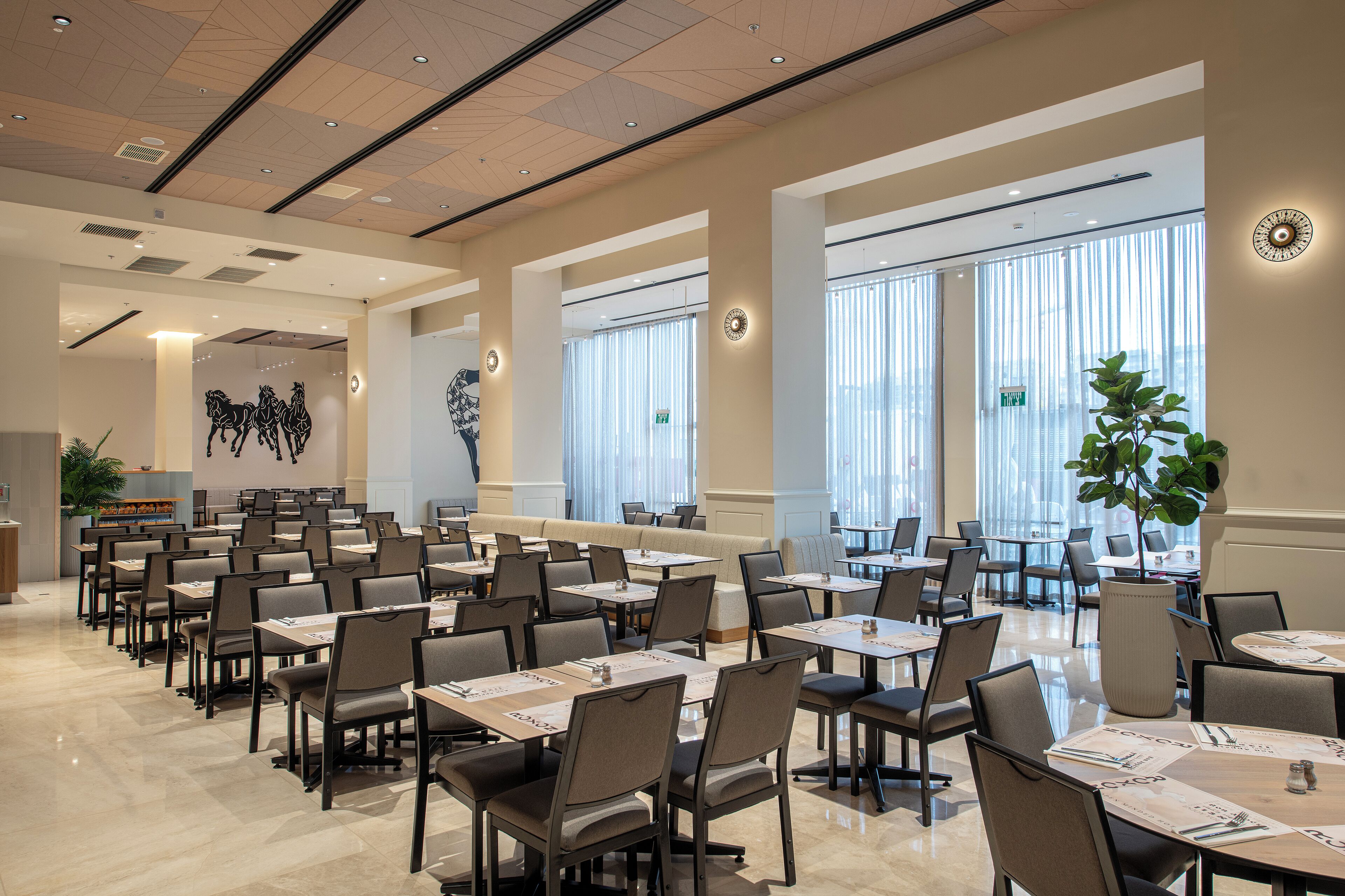 Un vaste espace dédié à la salle à manger, avec plusieurs tables et chaises, de grandes fenêtres équipées de stores et un élément décoratif mural.