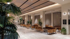 A spacious lobby with a long reception desk, multiple seating areas with sofas and armchairs, and a large potted plant in the foreground.