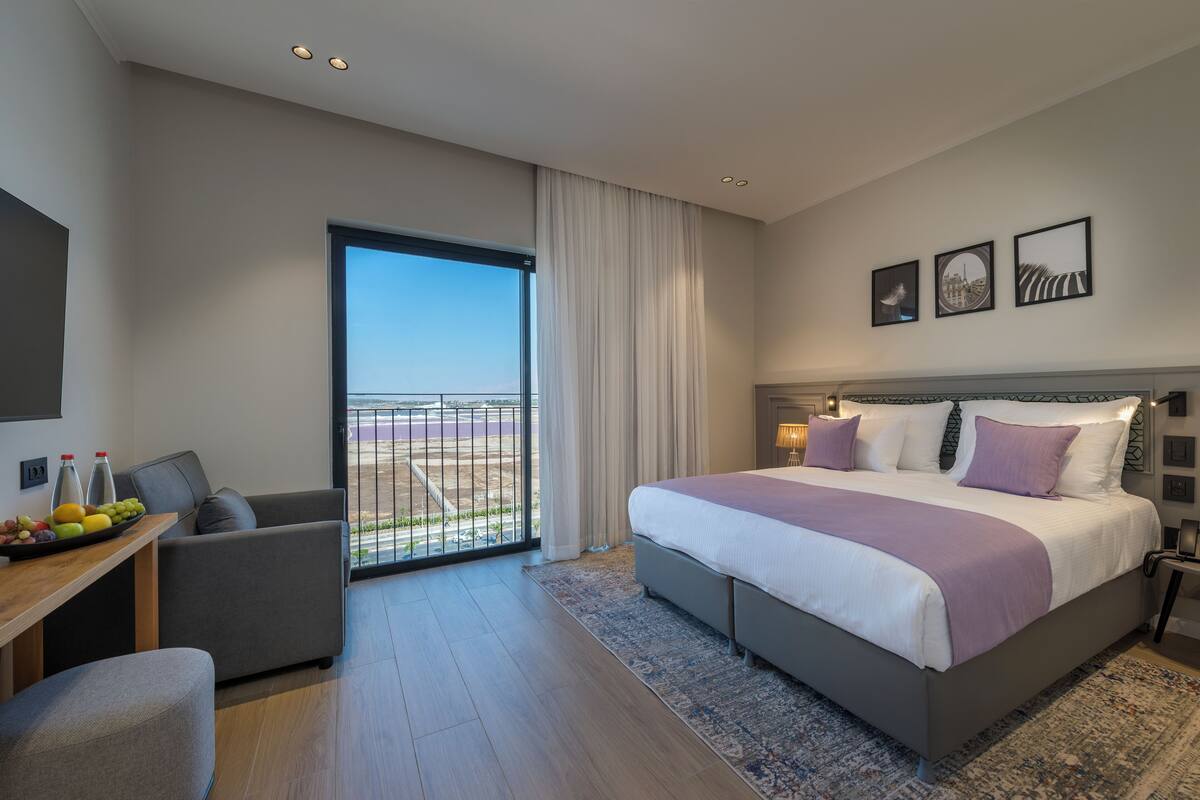 A modern hotel room with a large bed, a desk with a fruit tray, a sofa, and a balcony with a city view.