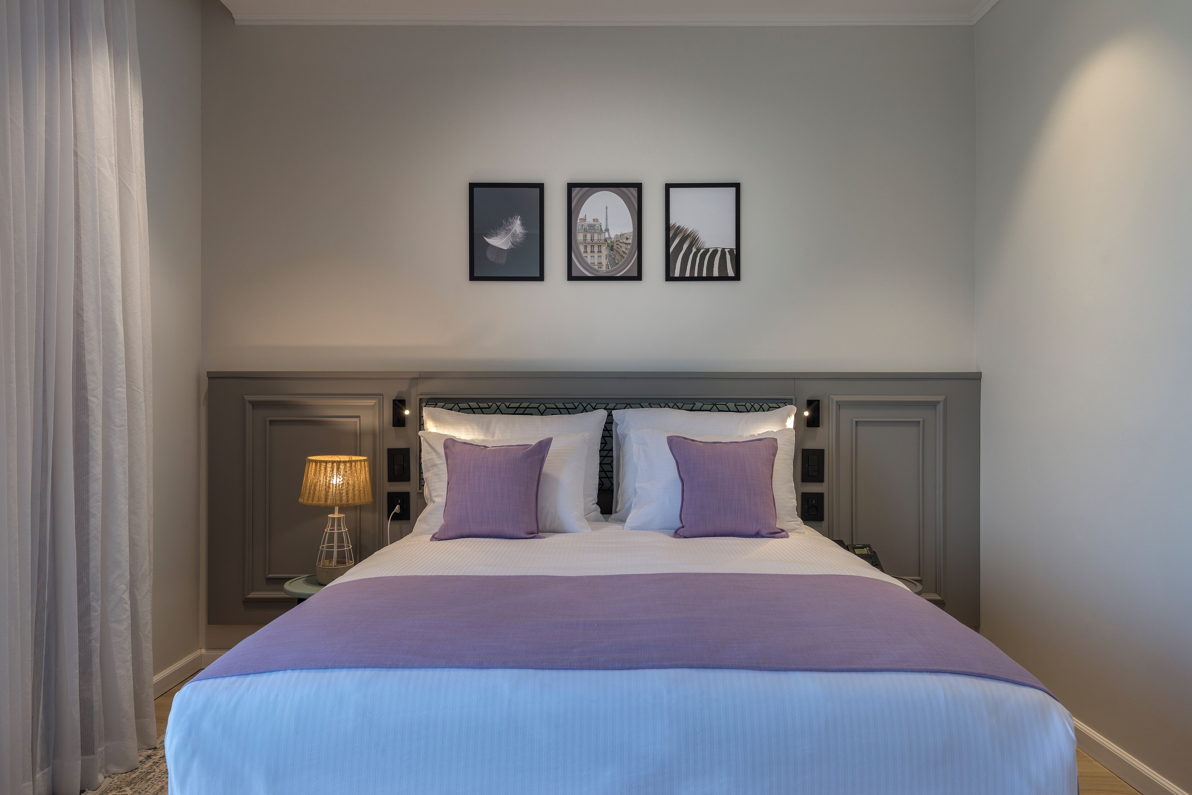 Un lit bien fait, recouvert d’une couverture violette, avec deux oreillers et une tête de lit ornée d’un motif géométrique. Trois tableaux encadrés sont accrochés au mur au-dessus du lit.