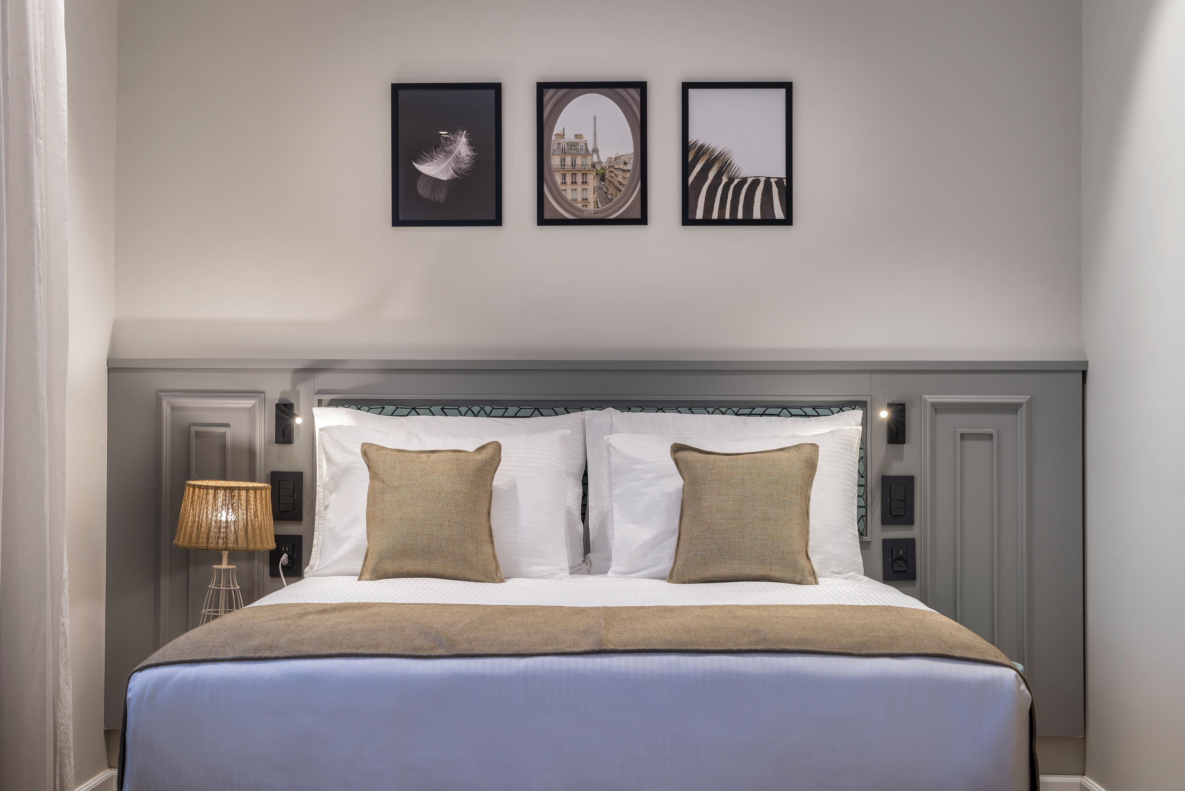 A neatly made bed with a white and blue bedspread, two beige pillows, and a headboard with a decorative trim. Above the bed, there are three framed pictures on the wall.