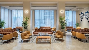 A spacious lobby with leather sofas, armchairs, and a coffee table. Large windows with sheer curtains allow natural light to fill the space.