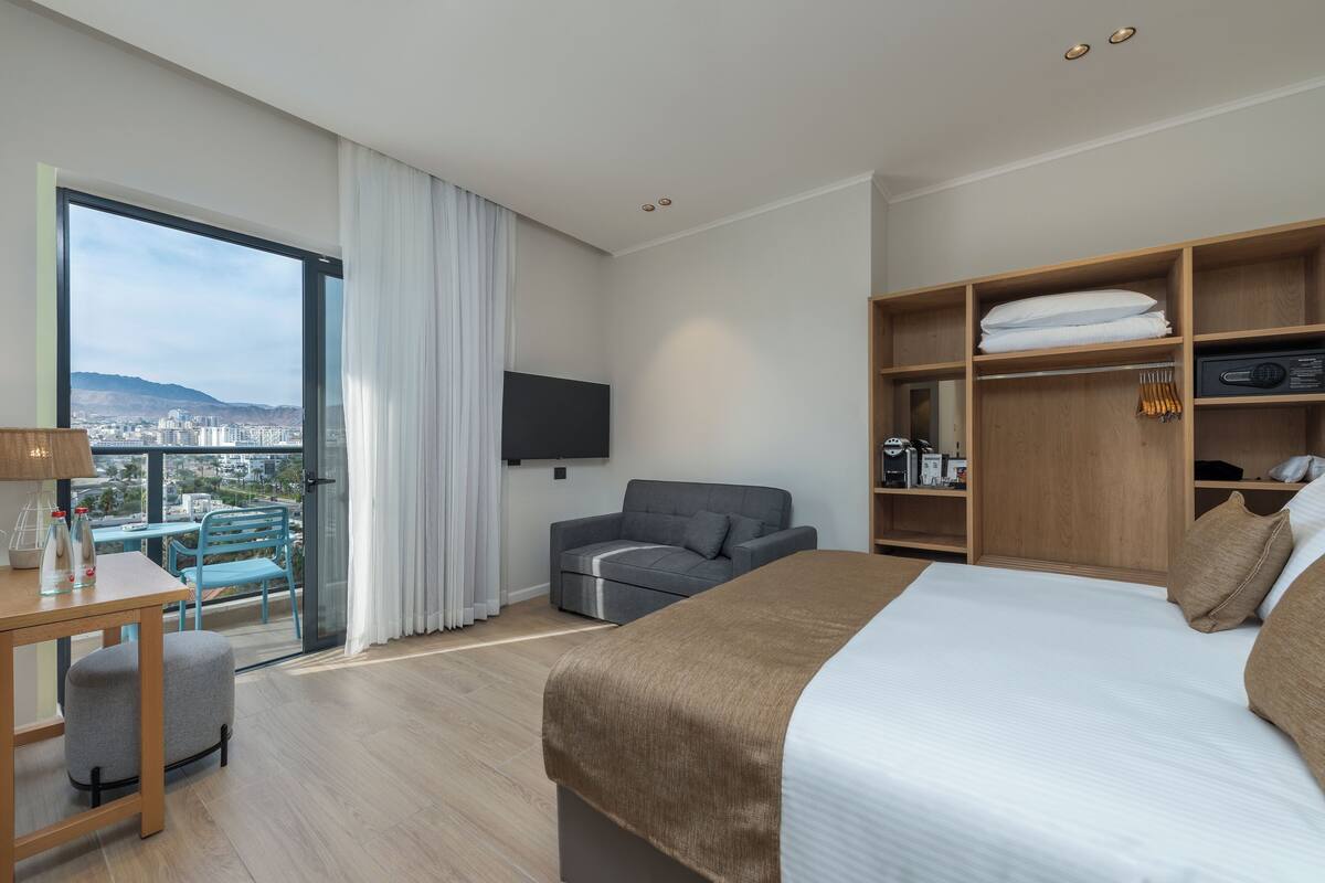 A modern hotel room with a large bed, a sofa, a TV, a wooden wardrobe, and a balcony with a city view.