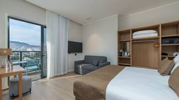 A modern hotel room with a large bed, a sofa, a TV, a wooden wardrobe, and a balcony with a city view.