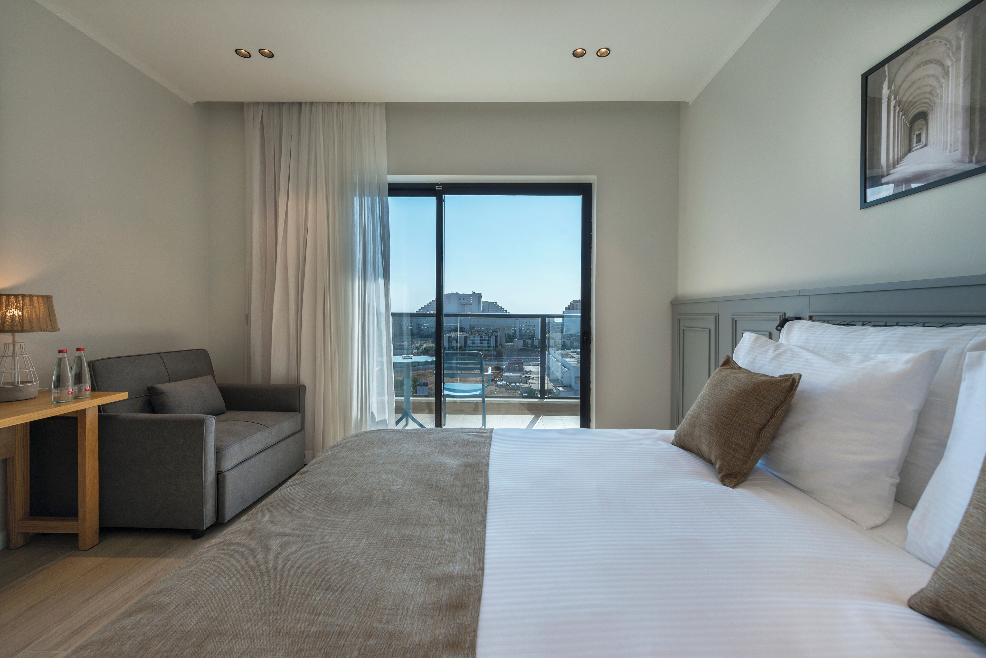 Une chambre d’hôtel avec un grand lit, un canapé, une petite table avec une lampe et une vue sur la ville, visible à travers une grande fenêtre.
