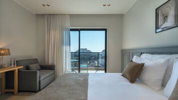 Ein Hotelzimmer mit einem großen Bett, einer Sitzecke, einem kleinen Tisch mit Lampe und Blick auf die Stadt durch ein großes Fenster.