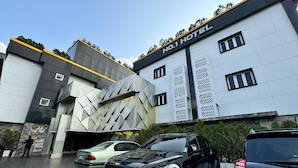 Un hotel moderno con una fachada geométrica llamativa, un estacionamiento con coches de lujo y un cielo despejado.