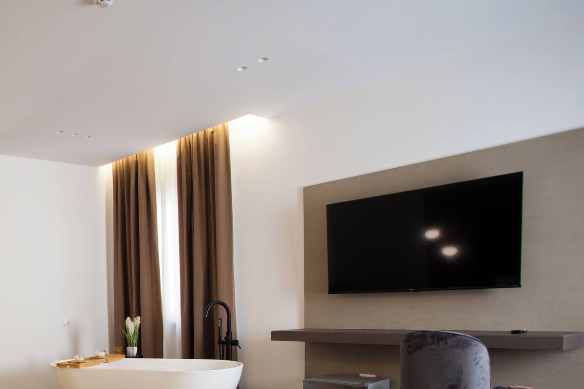 Ein modernes Hotelzimmer mit einem großen Bett, einer Badewanne, einem flachbildfernseher an der Wand und einem kleinen Schreibtisch mit Stuhl.