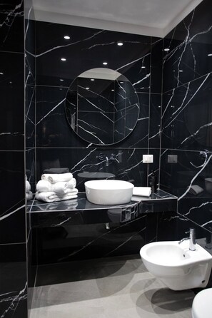 A modern bathroom with a black marble design, a round mirror, a white sink, and neatly folded towels. - Alexander House Hotel  (San Ferdinando)