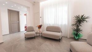 A modern hotel lobby with a neutral color scheme, featuring two sofas, a small table with a vase of flowers, and a potted plant. - Alexander House Hotel  (San Ferdinando)