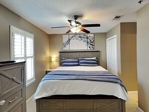 Une chambre à coucher comprenant un lit, une table de chevet, une commode, un ventilateur de plafond et des œuvres d’art accrochées au mur.