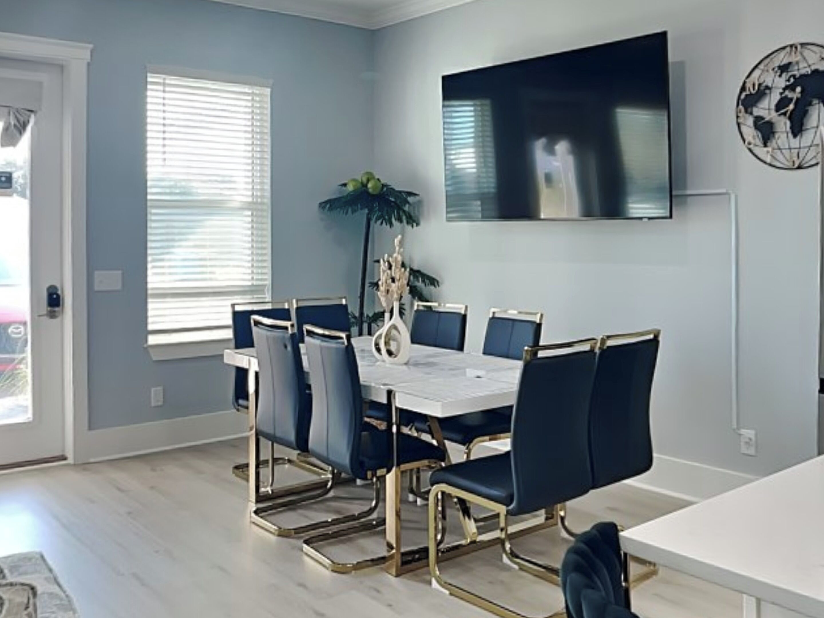 A dining area with a white table and blue chairs, a large flat-screen TV on the wall, and a potted plant.