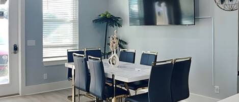 A dining area with a white table and blue chairs, a large flat-screen TV on the wall, and a potted plant.
