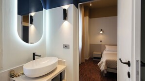 Un baño moderno con un lavabo ovalado grande, un espejo y una puerta que da a un dormitorio con cama y mesita de noche.