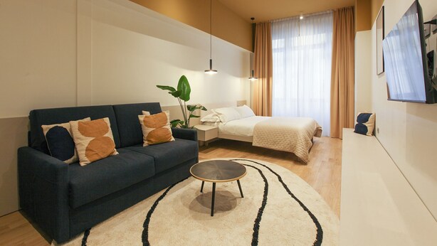 Una habitación de hotel moderna con un sofá azul oscuro, una mesa redonda de centro, una cama con edredón blanco y un amplio ventanal con cortinas de gasa.