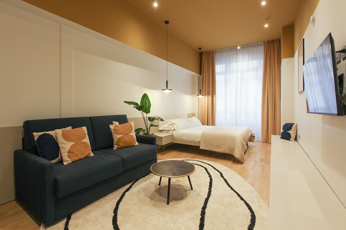 Une chambre d’hôtel moderne avec un canapé bleu foncé, une table basse ronde, un lit recouvert d’une couette blanche et une grande fenêtre avec des rideaux légers.
