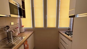 Une cuisine compacte dotée d’un four encastré, d’une cuisinière à gaz et d’une fenêtre avec des stores jaunes.