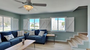 Ein großzügiges Wohnzimmer mit einem blauen Sofa, einem Holztisch und einem Deckenventilator. Große Fenster mit Fensterläden bieten Blick auf die Gebäude vor der Unterkunft.