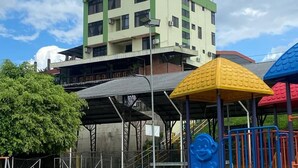 A playground with a slide and climbing structures, surrounded by a fenced area with a building in the background. - Hostal la Colina (Tena)