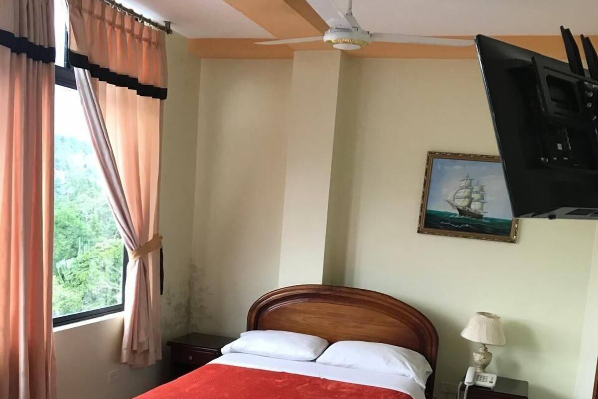 A bedroom with a bed, a nightstand, a desk, a ceiling fan, a TV mounted on the wall, and a window with curtains.