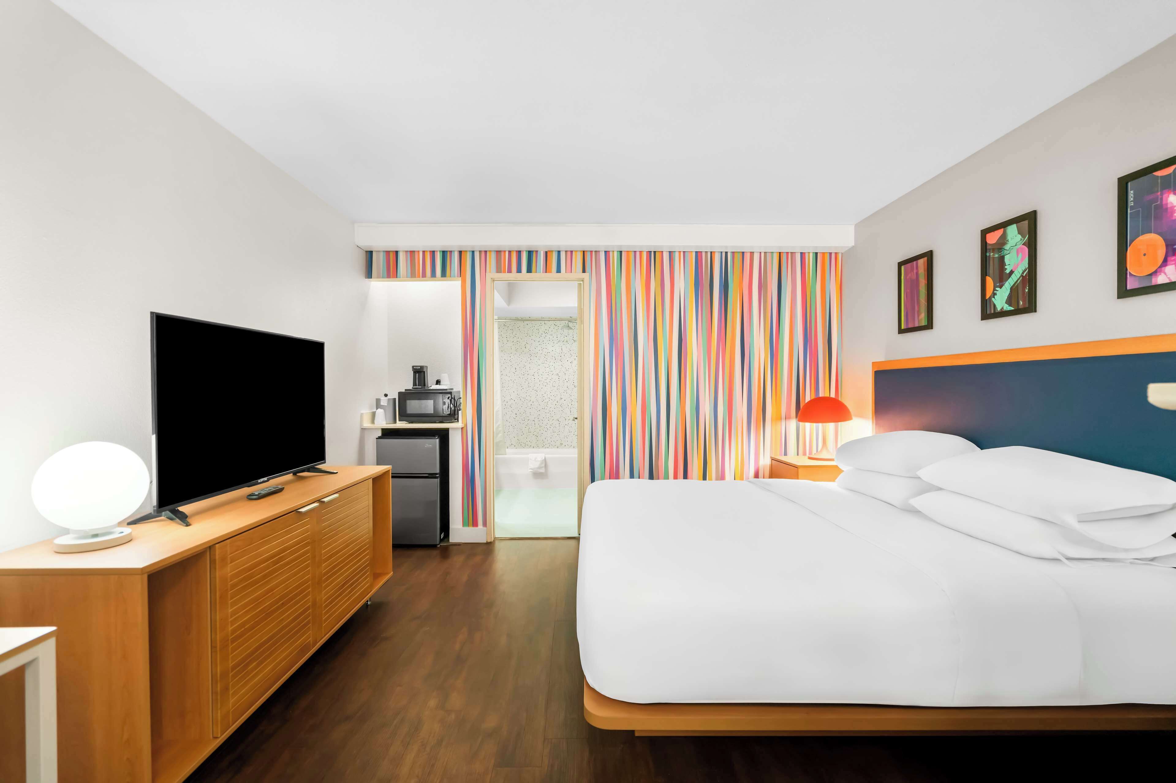 A hotel room with a bed, a TV, a desk with a lamp, and a bathroom visible through a door.