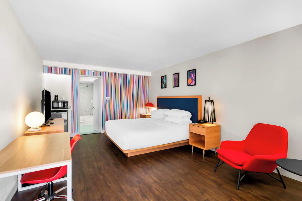 A modern hotel room with a wooden bed, a desk with a lamp, a red chair, and a TV.