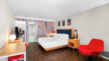 A modern hotel room with a wooden bed, a desk with a lamp, a red chair, and a TV.