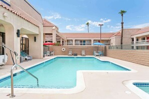 A swimming pool with lounge chairs and umbrellas, surrounded by a building with a tiled roof and palm trees.