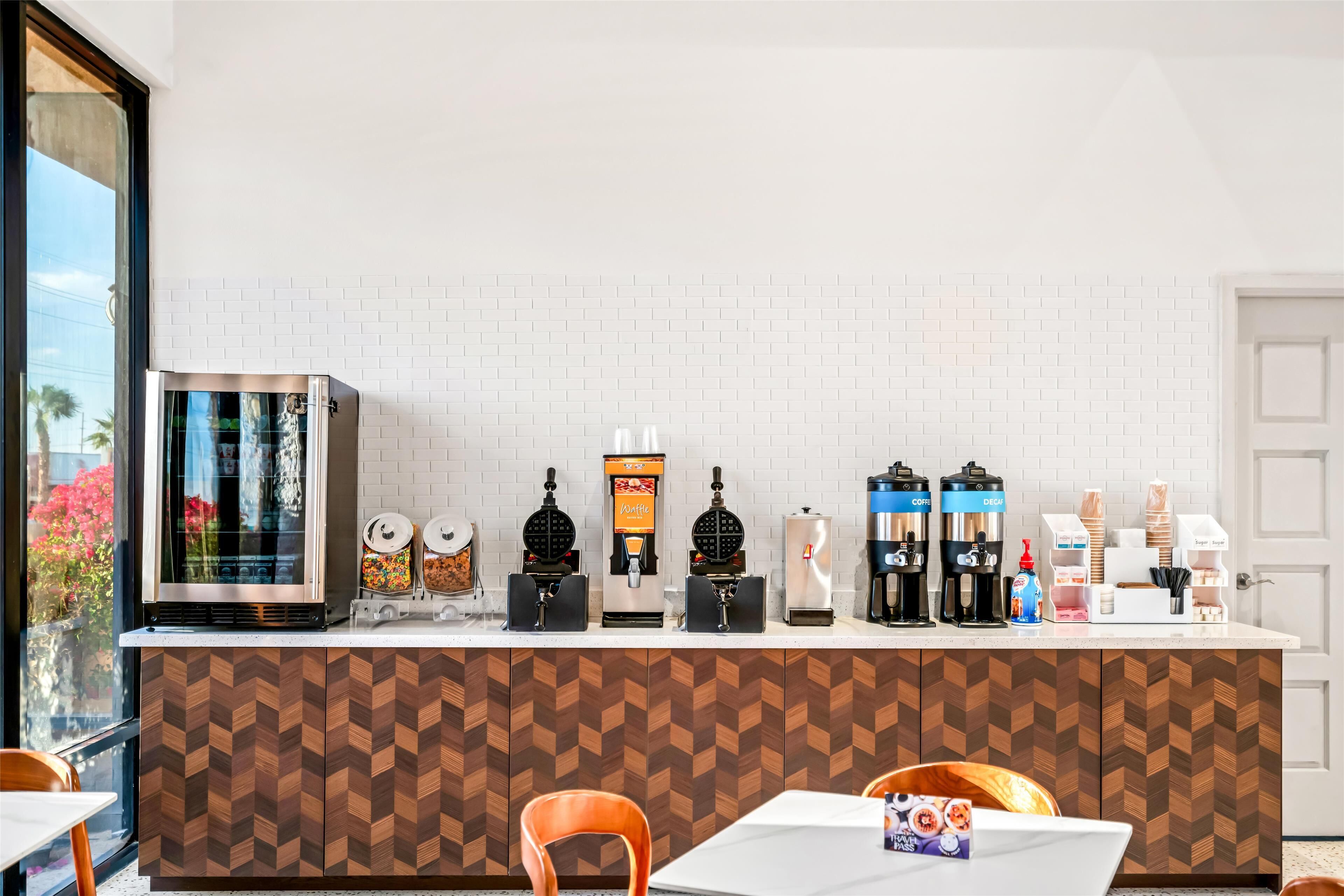 Een moderne koffiebar met diverse koffieapparaten, een gaufre apparaat en een koelkast. Er staan stoelen en tafels op de voorgrond.