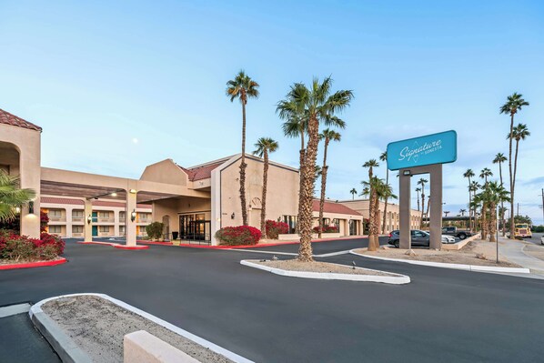 A hotel entrance with a sign, palm trees, and a parking lot. - Signature Indio I-10 Coachella Valley by Sonesta (Indio)