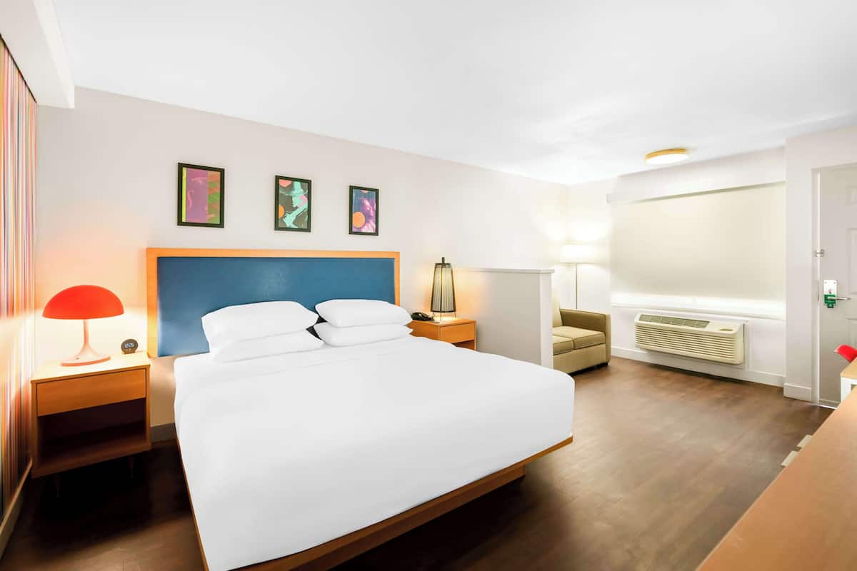 A hotel room with a large bed, a bedside table with a lamp, a chair, and a wall-mounted air conditioner.