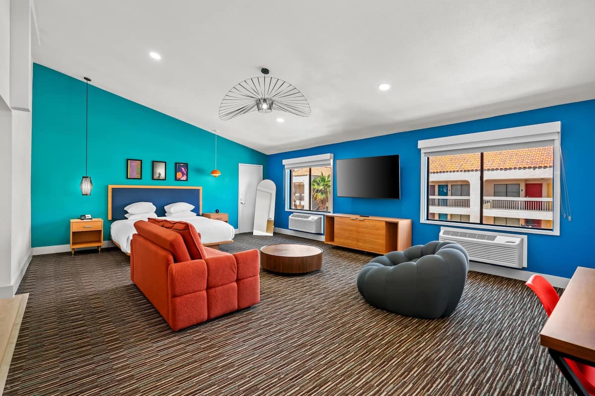 A modern hotel room with a teal accent wall, a large bed, a TV, a desk, and a window with a view of the outside.