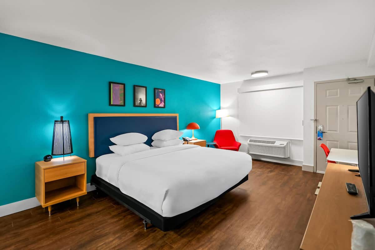 A modern hotel room with a large bed, a nightstand, a red chair, a desk with a TV, and a teal-colored wall.