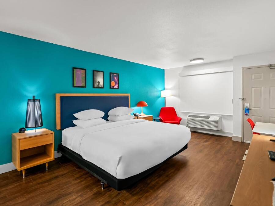 Ein modernes Hotelzimmer mit einem großen Bett, einem Nachttisch, einem roten Sessel, einem Schreibtisch mit Fernseher und einer türkisblauen Wand.