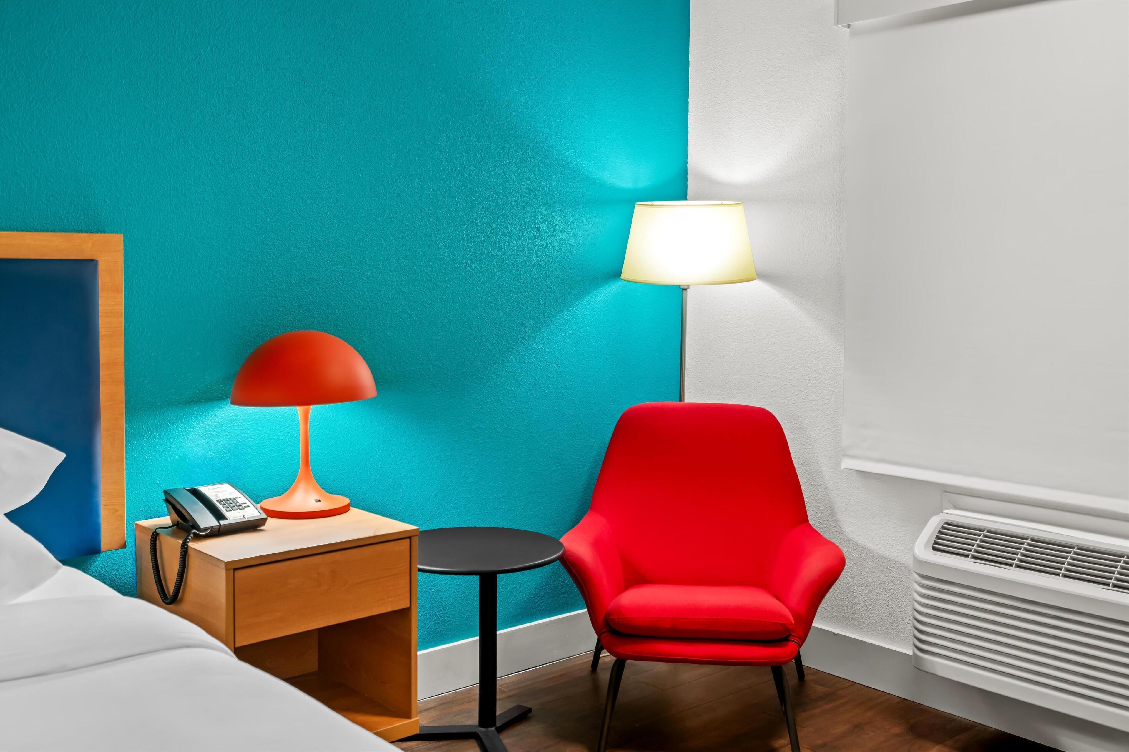 Une chambre d’hôtel avec un mur de couleur turquoise, une chaise rouge, une petite table en bois avec une lampe, un téléphone sur la table de chevet et un radiateur blanc.