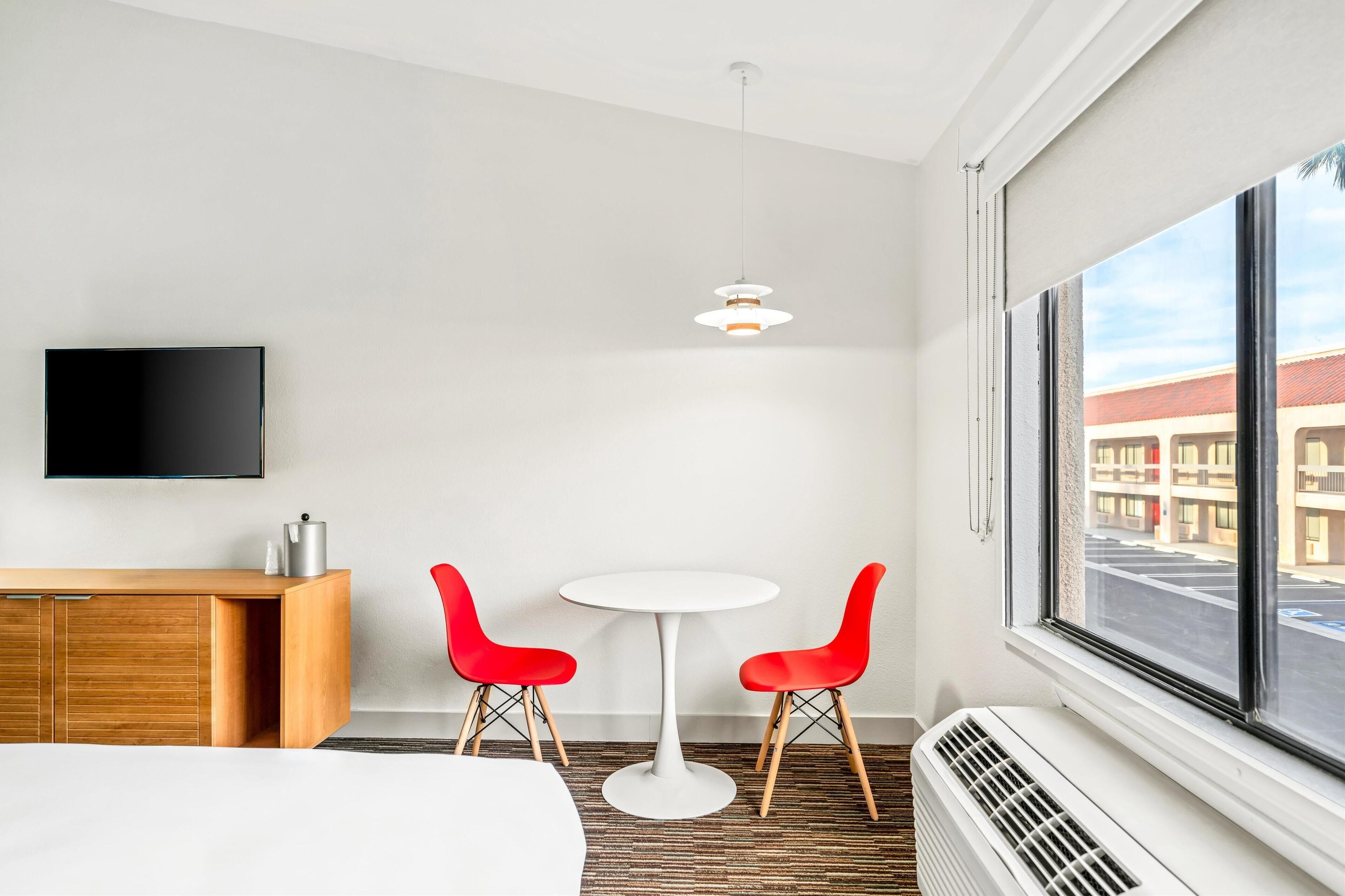 Habitación de hotel con cama, televisor, una pequeña mesa con dos sillas rojas, una lámpara colgante blanca y un ventanal con vistas a edificios.