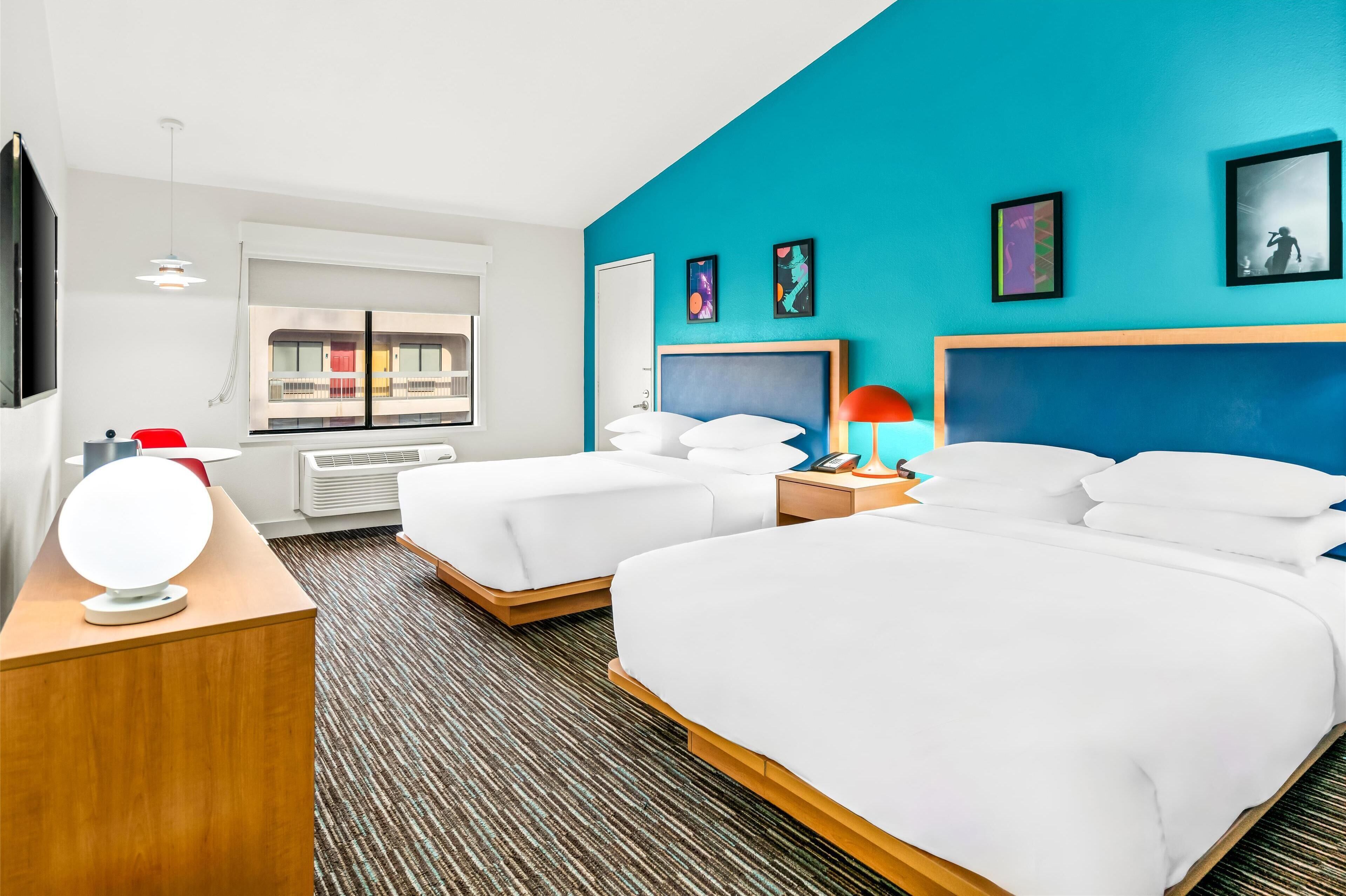 A hotel room with two beds, a TV, a window with a view of a balcony, and artwork on the walls.