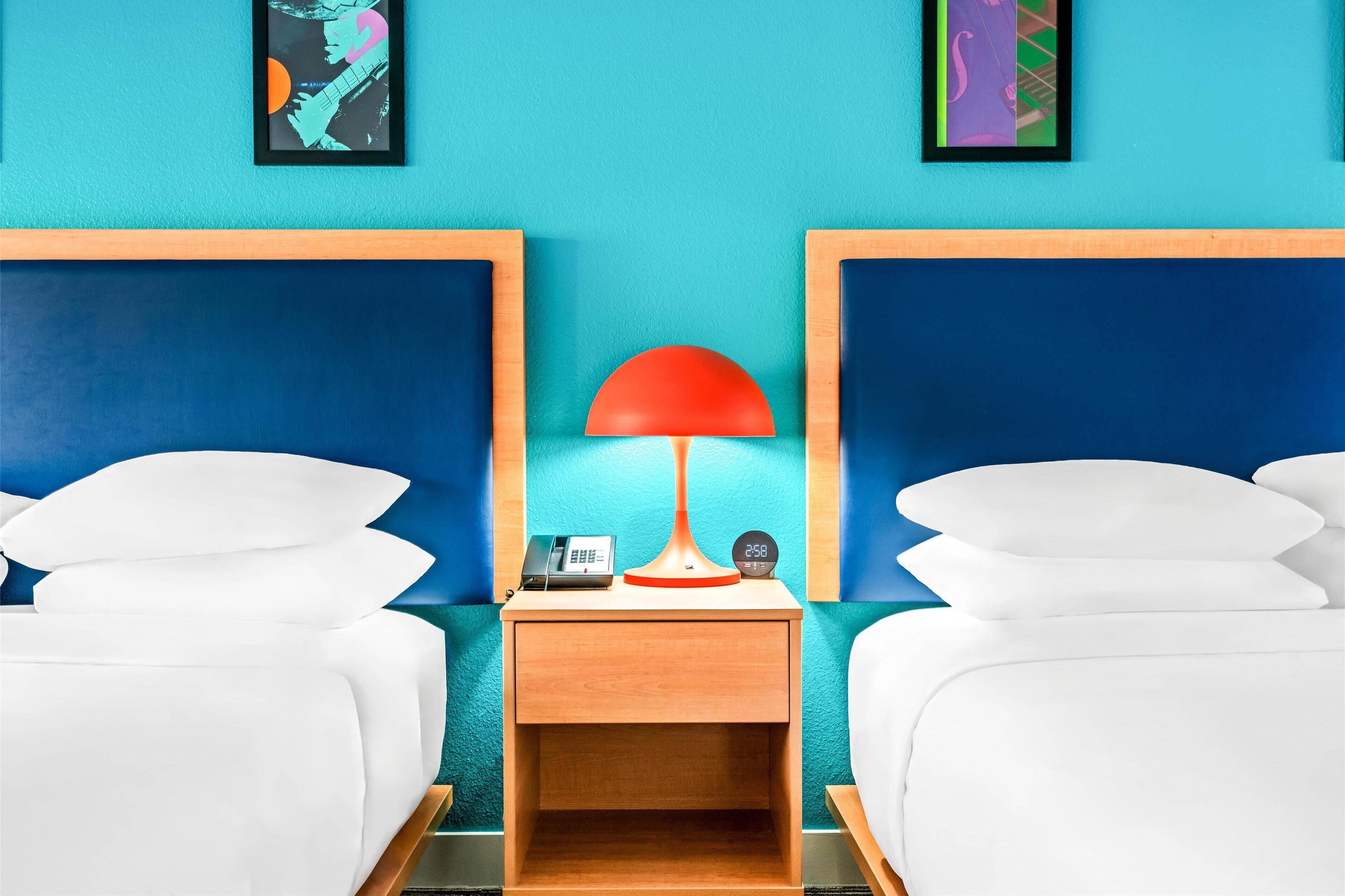 A hotel room with two beds, a nightstand with a lamp, a phone, and a clock.