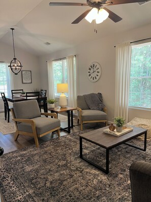 Un salon doté d’un ventilateur de plafond, d’une table à manger avec des chaises, d’une table basse agrémentée d’objets décoratifs et d’une grande fenêtre avec des rideaux.