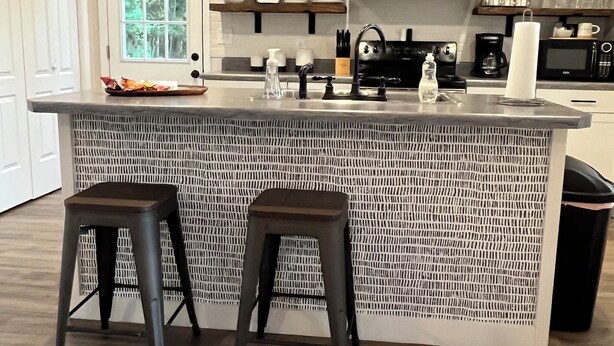 Une cuisine moderne dotée d’un îlot central, de deux tabourets, d’une hotte noire et d’étagères en bois au-dessus du plan de travail.