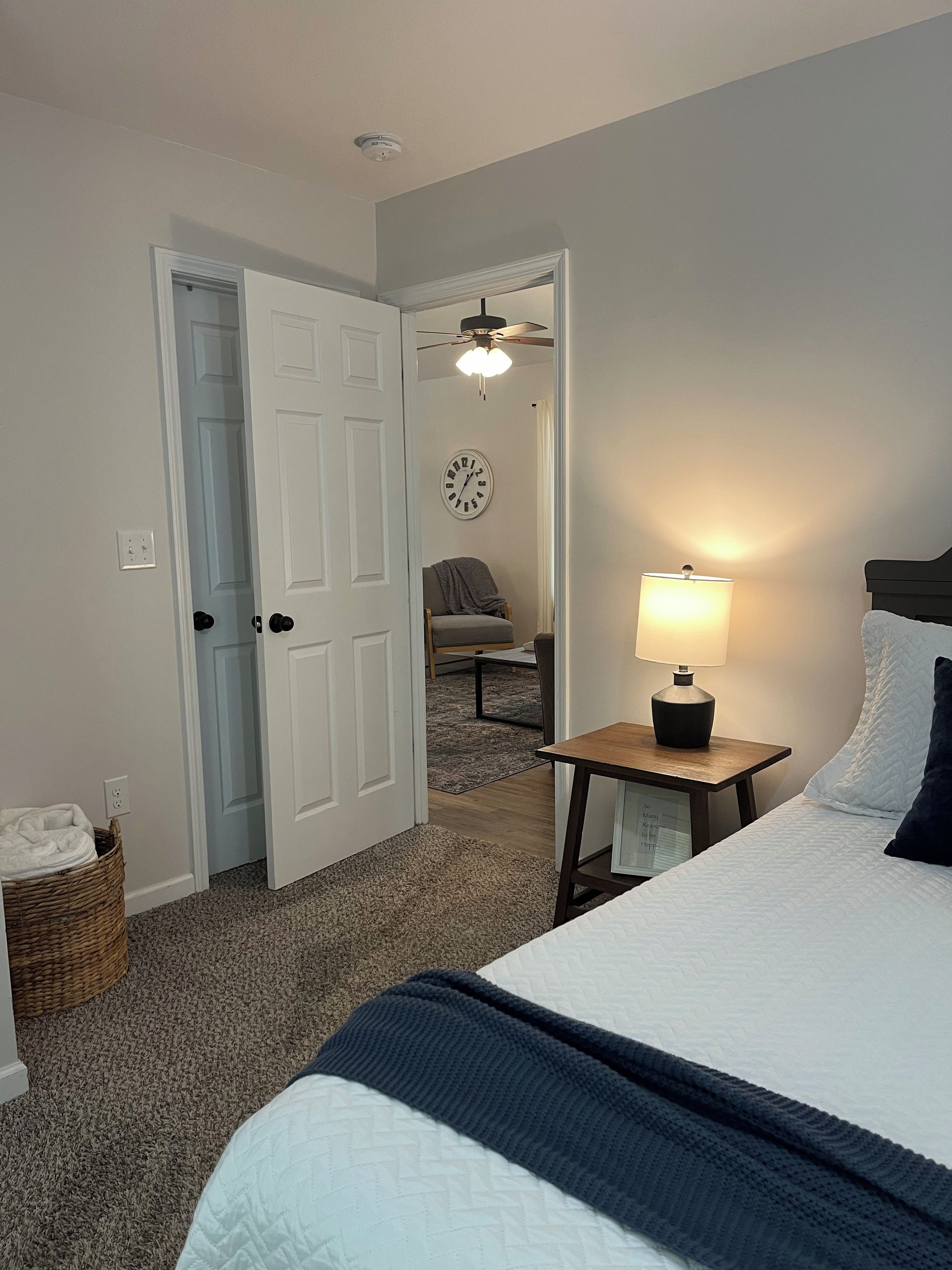 Habitación de hotel con cama, mesita de noche con lámpara, reloj de pared, ventilador de techo y una puerta que da a otra habitación.