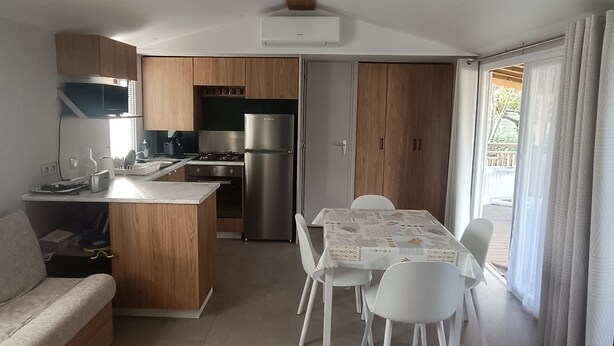 Une cuisine moderne avec des armoires en bois, une cuisinière encastrée et un réfrigérateur en acier inoxydable. On y trouve un coin repas avec une table et des chaises, ainsi qu’un canapé dans l’espace salon.
