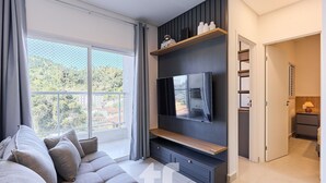 Una sala de estar moderna con un ventanal grande, un televisor de pantalla plana montado en una pared oscura, un sofá y una estantería de madera con objetos decorativos.