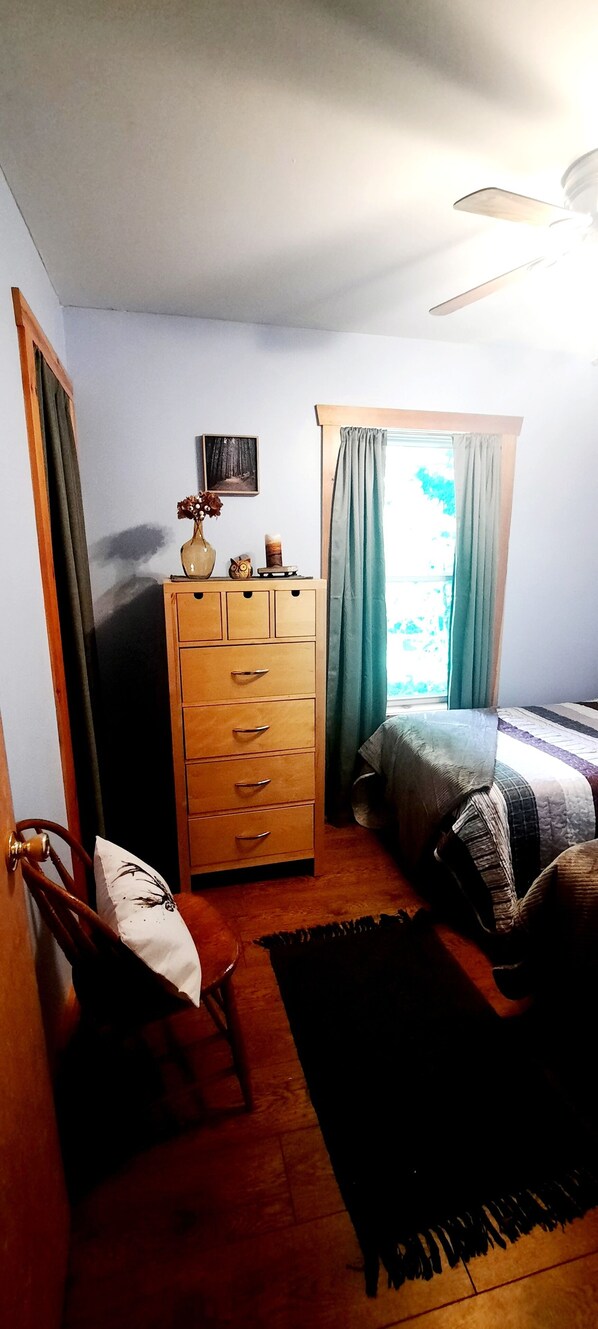 A bedroom with a wooden dresser, a bed with a striped blanket, a chair, and a window with curtains.